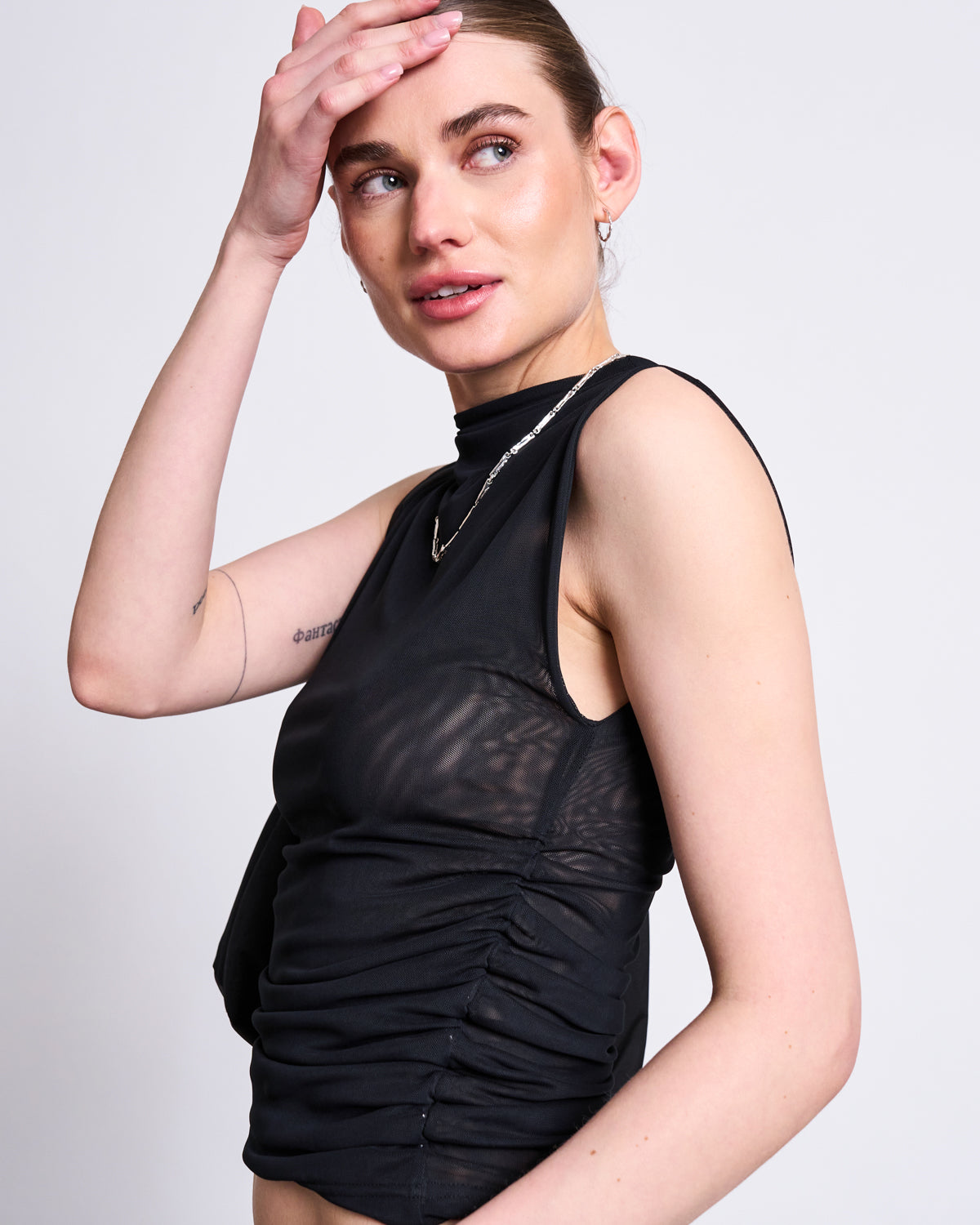 A woman wearing the jan-n-june RUFFLE MESH TOP LAGUNA BLACK poses with one hand touching her forehead, gazing slightly upward to the side against a plain light backdrop.