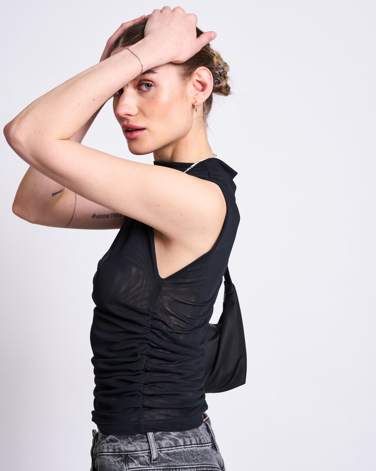 Wearing the jan-n-june RUFFLE MESH TOP LAGUNA BLACK and gray jeans, a person strikes a pose with both hands on their head, gazing slightly aside. They carry a small black shoulder bag against a plain light gray background.