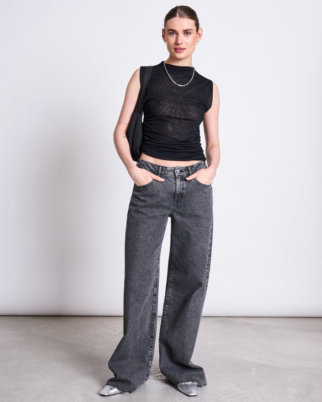 A woman stands against a plain light background, hands in her pockets and exuding confidence, wearing the jan-n-june RUFFLE MESH TOP LAGUNA BLACK, loose gray jeans, silver pointed shoes, a necklace, and a black shoulder bag.