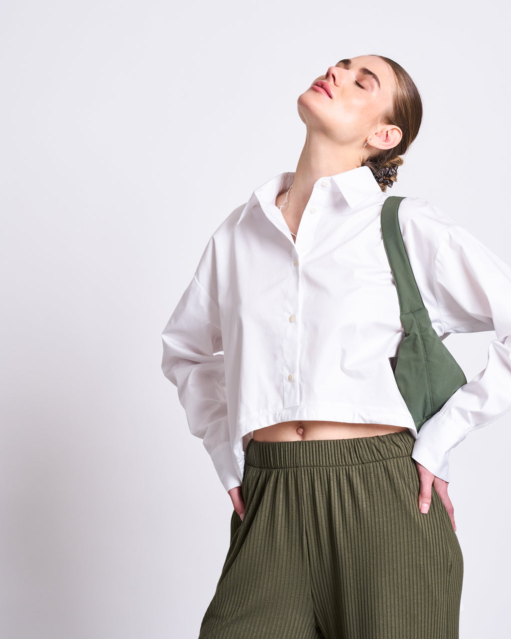 A woman wears the jan-n-june DRAWSTRING SHIRT ISTED WHITE, a 100% cotton oversized button-up, paired with green pleated pants and a green shoulder bag. She stands against a neutral background, hand on hip, head tilted back, eyes closed.