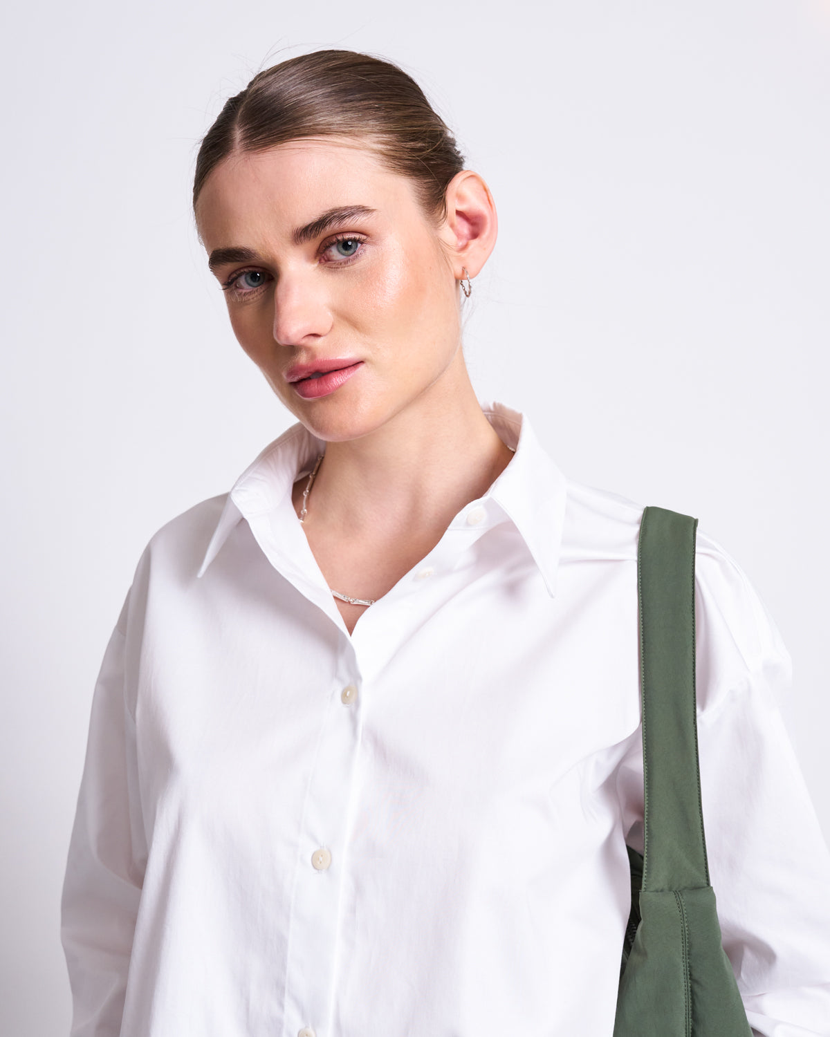 A woman with light skin and blue eyes wears the jan-n-june DRAWSTRING SHIRT ISTED WHITE, featuring an oversized fit and dropped shoulders. She stands against a plain background, accessorized with small hoop earrings and a green bag strap.