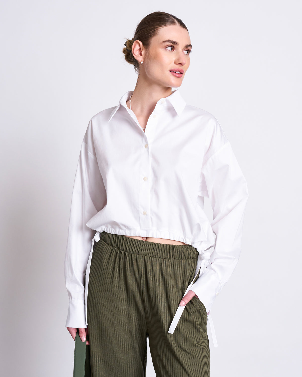 A woman with brown hair in a bun wears the jan-n-june DRAWSTRING SHIRT ISTED WHITE, a loose 100% cotton button-up with dropped shoulders, paired with green pleated pants. She stands against a white background, looking right with one hand in her pocket.