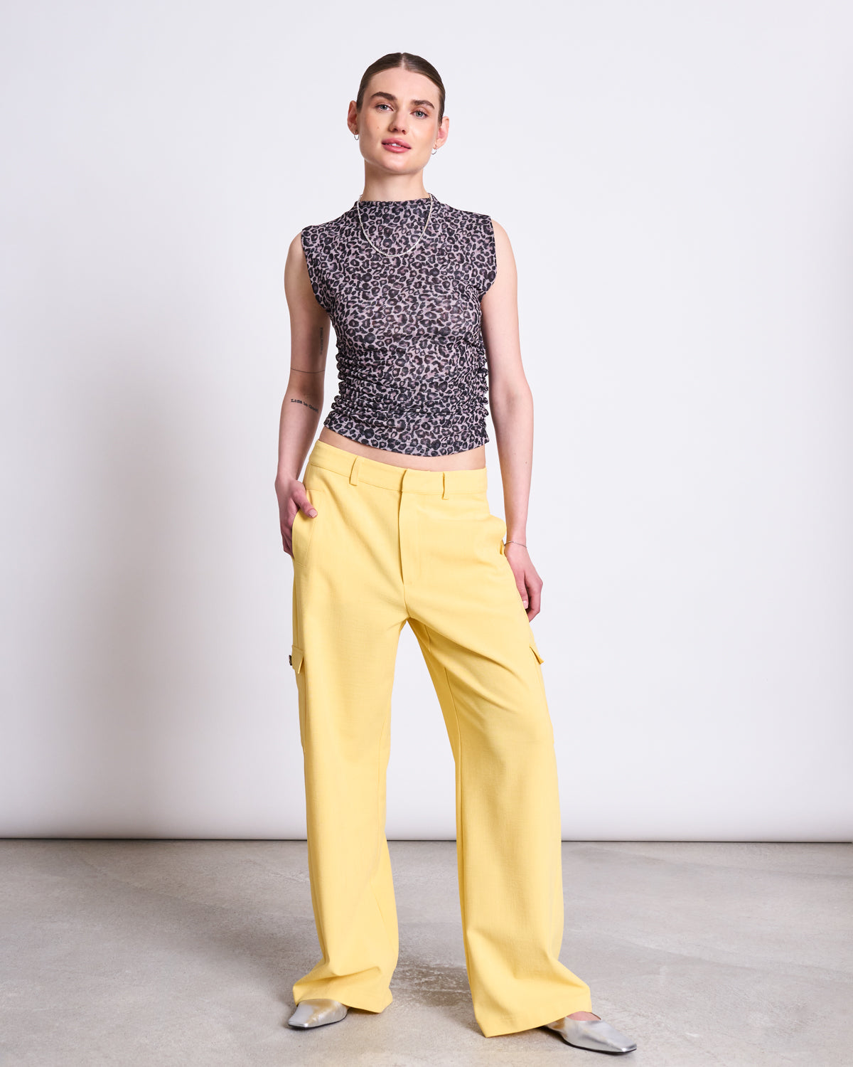 A person stands against a plain white wall wearing the jan-n-june RUFFLE TOP LAGUNA LEO PRINT, a sleeveless black and grey patterned sheer top, paired with loose yellow pants with side pockets and silver shoes. Hands are in pockets, facing the camera.