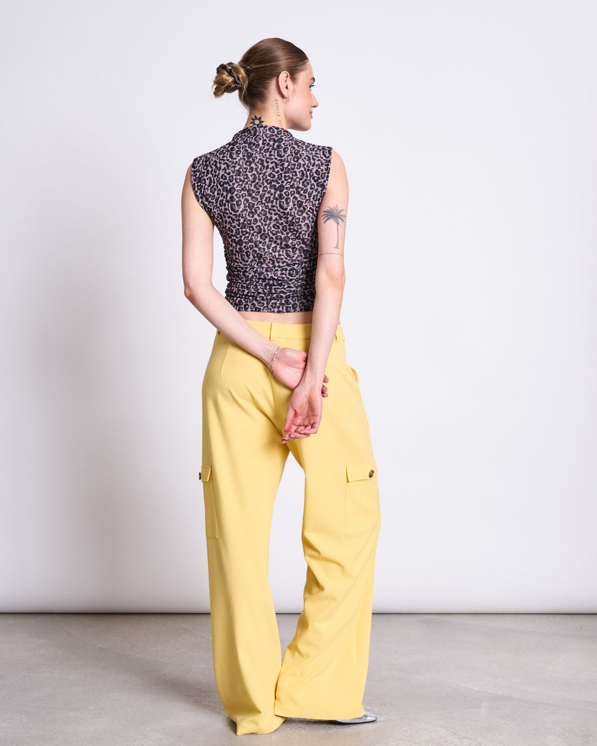 A woman with her hair in a bun, hands behind her back, wears the jan-n-june RUFFLE TOP LAGUNA LEO PRINT—a sleeveless, high-neck leopard mesh top—with loose yellow pants against a plain light background.