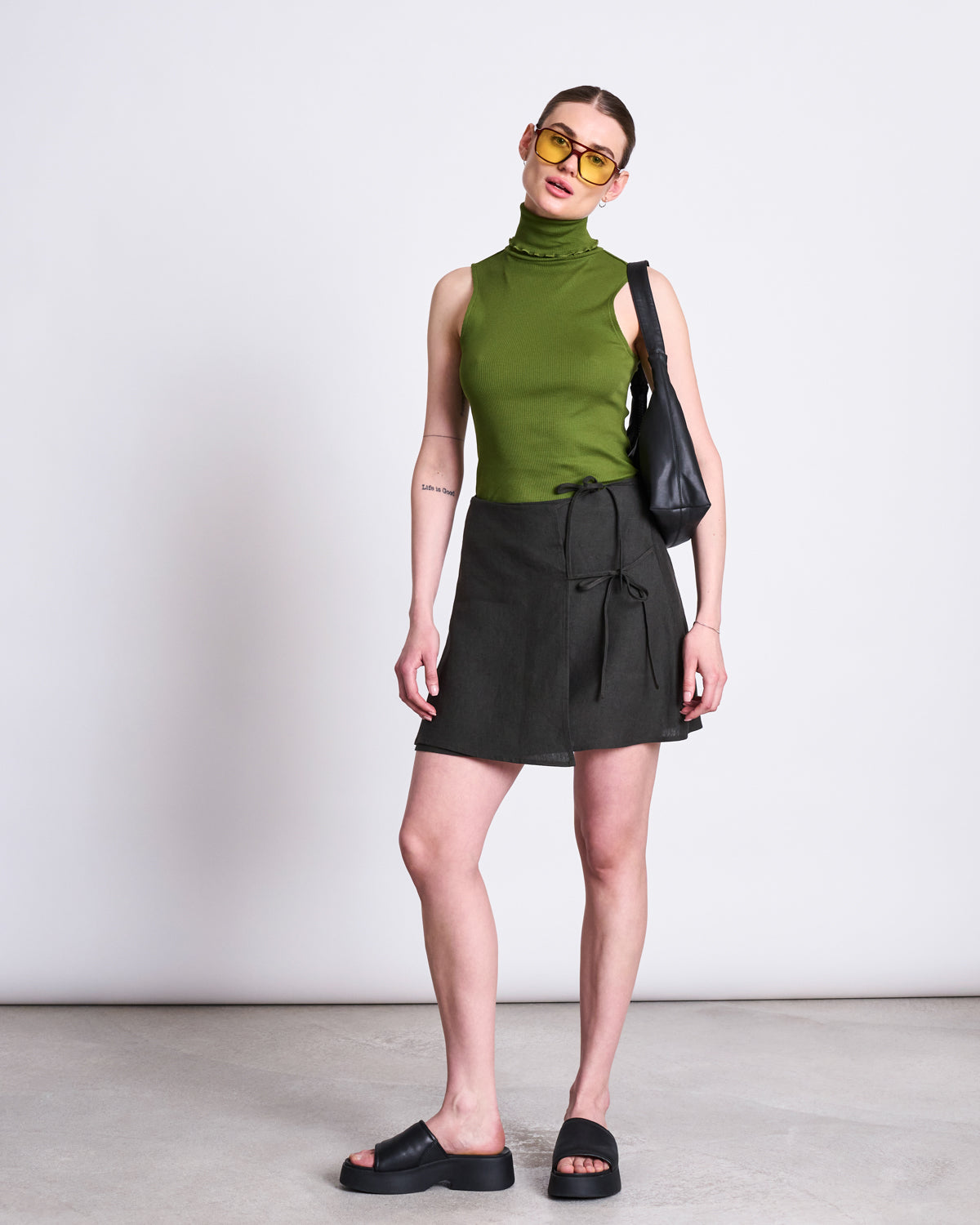 A woman wearing yellow sunglasses, a sleeveless green turtleneck, the jan-n-june LINEN TIE SKIRT KYS DARK OLIVE, black slide sandals, and a black shoulder bag stands against a plain white background.