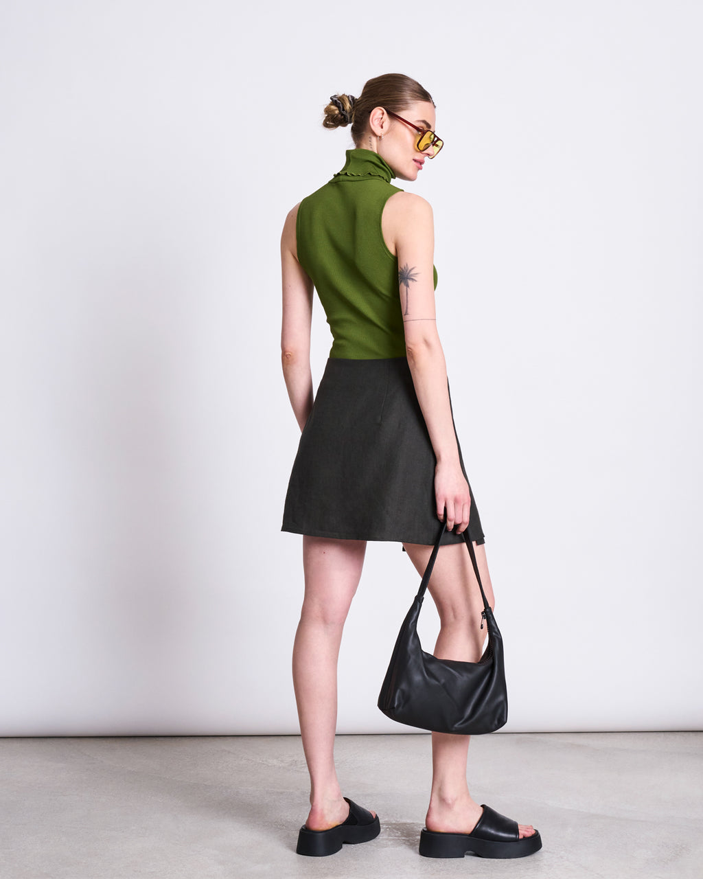 A woman stands facing away, wearing a sleeveless green turtleneck, black sandals, sunglasses, and her hair in a bun. She holds a black handbag and wears the jan-n-june LINEN TIE SKIRT KYS DARK OLIVE. The background is plain and light-colored.
