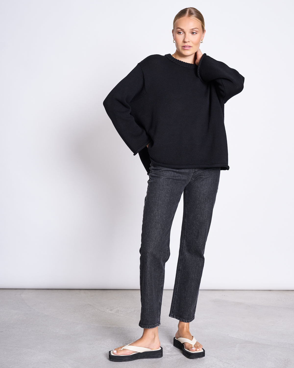 A person stands against a plain background wearing the jan-n-june JUMPER SONA BLACK GOTS, an oversized organic cotton knit sweater, paired with dark straight-leg jeans and white sandals. One hand touches their neck, the other rests by their side.