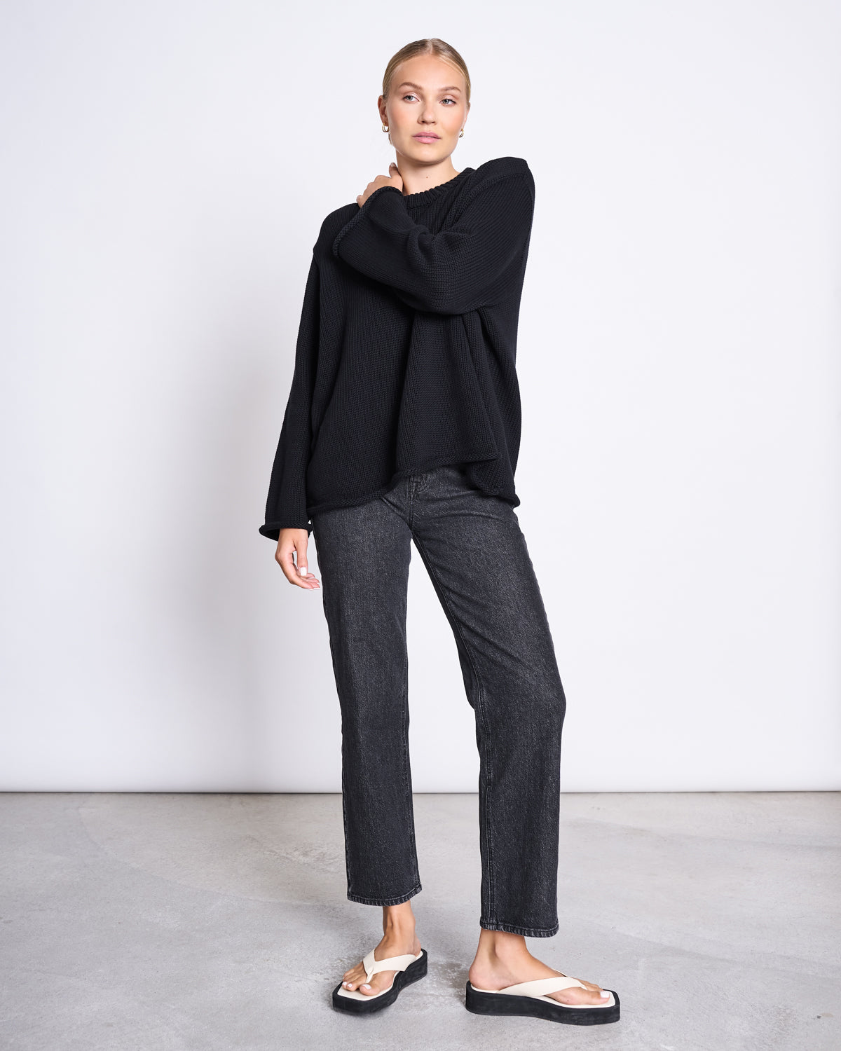 A woman with blonde hair tied back stands against a plain background wearing the jan-n-june JUMPER SONA BLACK GOTS, an oversized organic cotton knit sweater, paired with dark gray straight-leg jeans and white flip-flop sandals. One hand rests on her opposite shoulder.