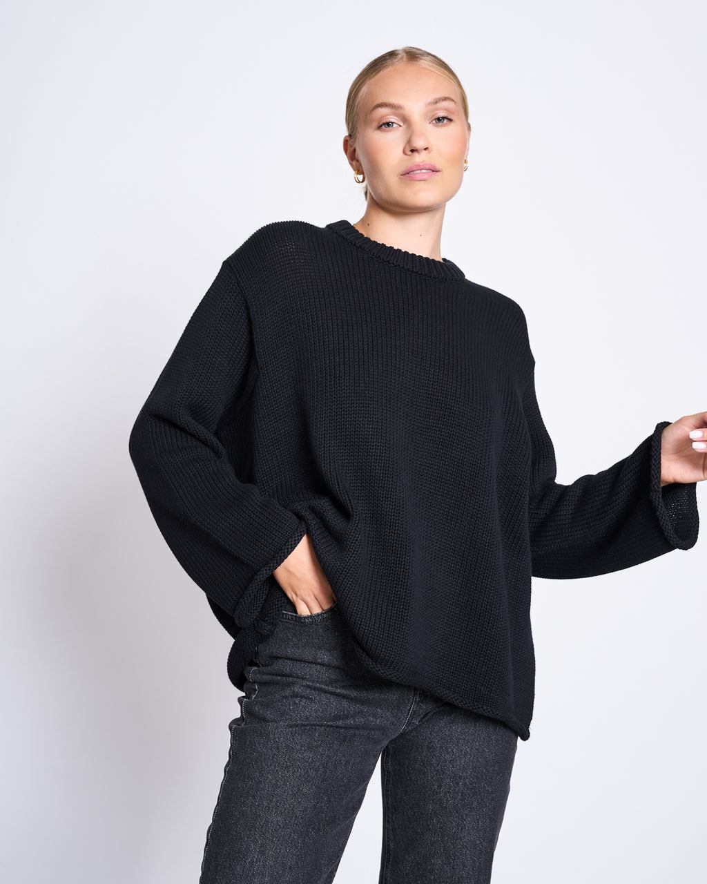 A woman with blonde hair in a neat bun wears the jan-n-june JUMPER SONA BLACK GOTS, an oversized knit black sweater made from GOTS certified organic cotton, paired with dark jeans, posing with one hand in her pocket against a white background.