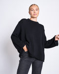 A woman with blonde hair in a neat bun wears the jan-n-june JUMPER SONA BLACK GOTS, an oversized knit black sweater made from GOTS certified organic cotton, paired with dark jeans, posing with one hand in her pocket against a white background.