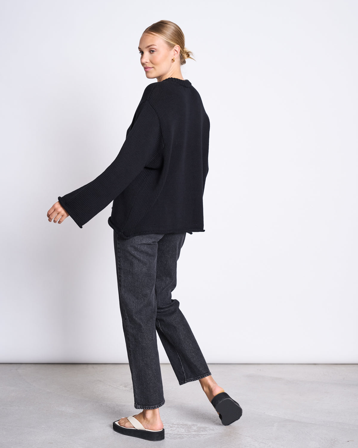A woman with blonde hair in a low bun wears the jan-n-june JUMPER SONA BLACK GOTS—an oversized organic cotton sweater—paired with dark straight-leg jeans and black platform sandals. She stands sideways, looking over her shoulder against a white background.