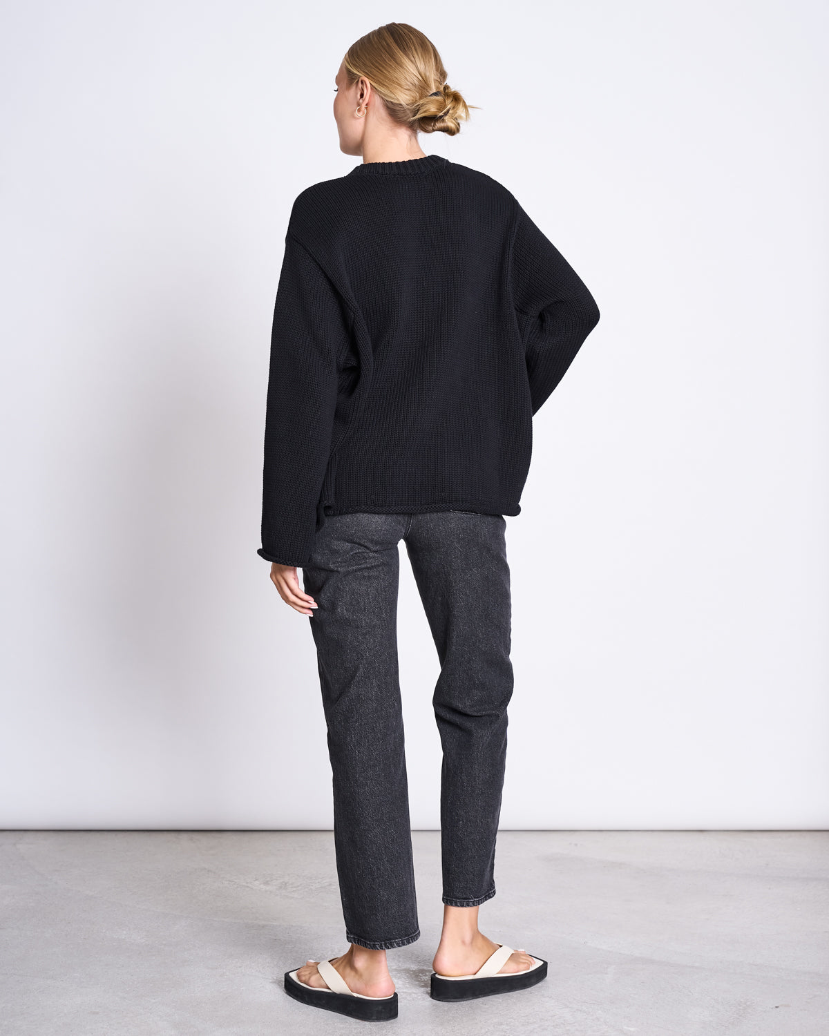 A person with blonde hair in a low bun stands facing away, wearing the jan-n-june JUMPER SONA BLACK GOTS—an oversized organic cotton knit sweater—with dark jeans and white slide sandals, against a plain white wall and gray floor.