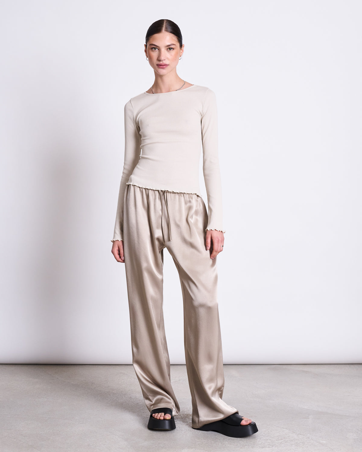 A woman stands against a plain white background wearing the jan-n-june MINIRIB LONGSLEEVE AVERSA IVORY GOTS, styled with loose satin beige pants and black platform sandals. She looks forward with her hair tied back.