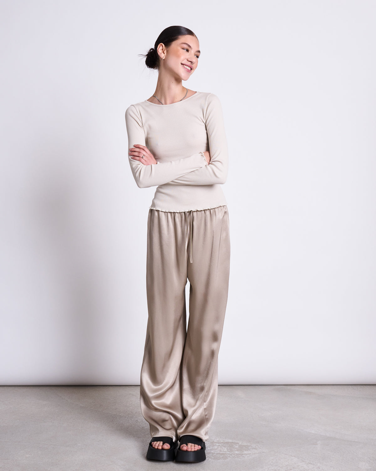 A woman smiles with arms crossed, wearing the jan-n-june MINIRIB LONGSLEEVE AVERSA IVORY GOTS top, loose satin taupe pants, and black slide sandals against a plain white background.