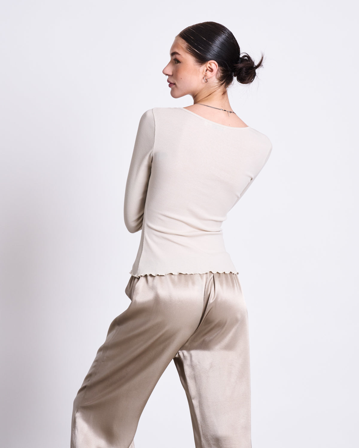 A woman with dark hair in a low bun stands sideways, wearing the jan-n-june MINIRIB LONGSLEEVE AVERSA IVORY GOTS top and shiny beige satin pants against a plain white background.