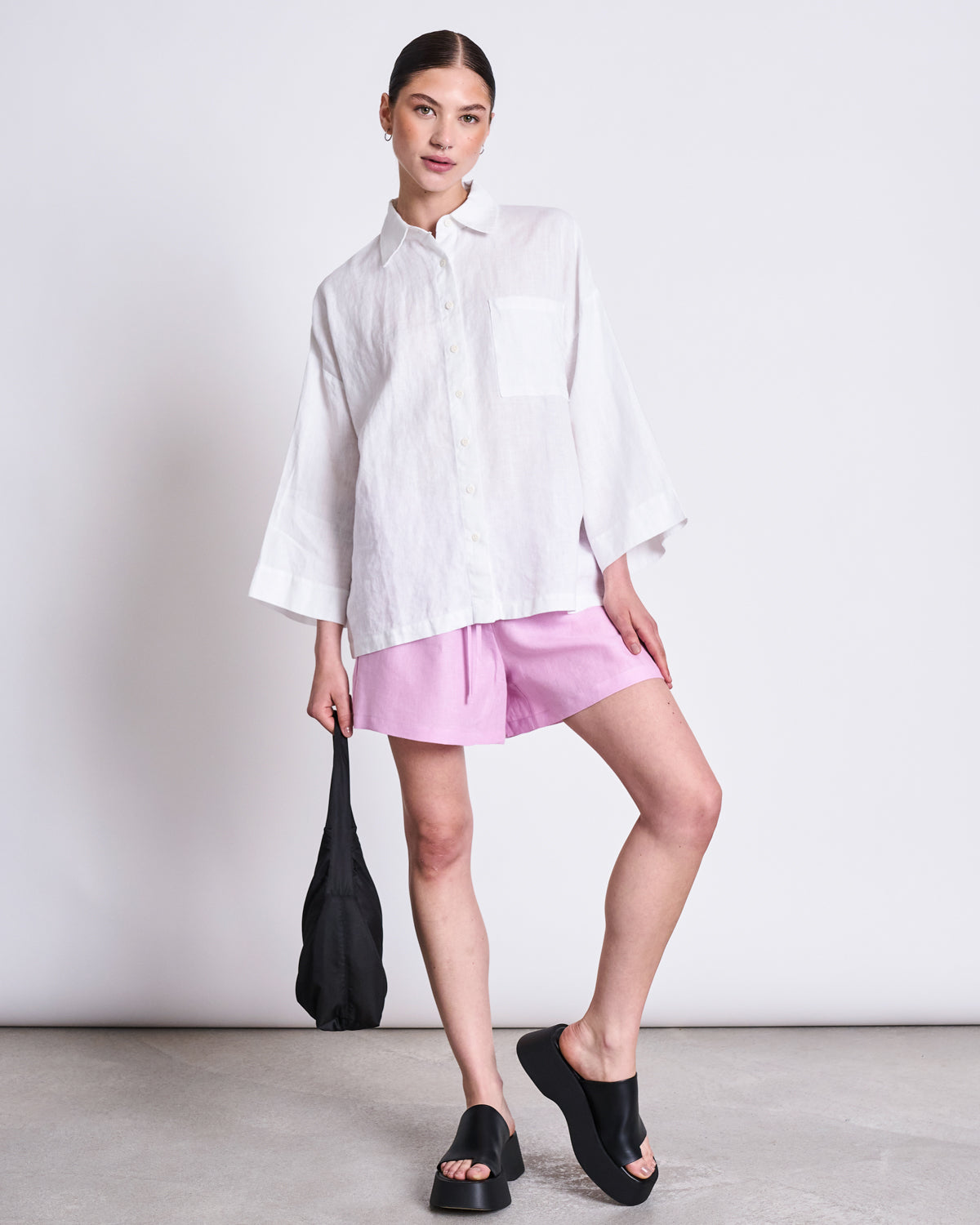 A woman stands against a plain background wearing the jan-n-june LINEN SHIRT MONS WHITE, styled with light pink shorts and black slide sandals. She holds a small black bag and gazes confidently at the camera.