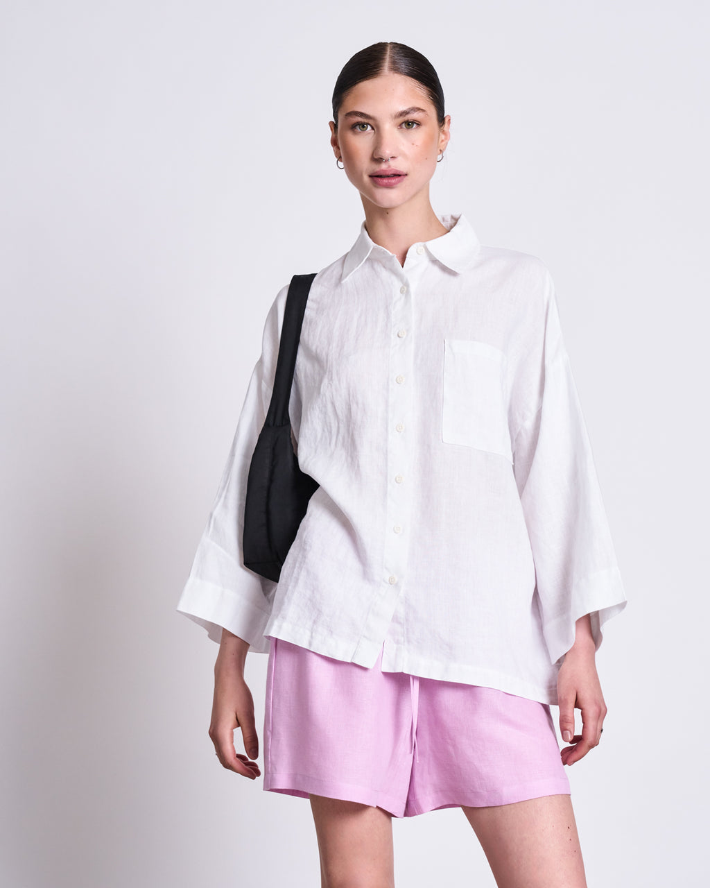 A woman stands against a plain background wearing the jan-n-june LINEN SHIRT MONS WHITE. She pairs the loose, classic-collar shirt with light pink shorts and a black shoulder bag, her hair tied back and her expression relaxed and confident.