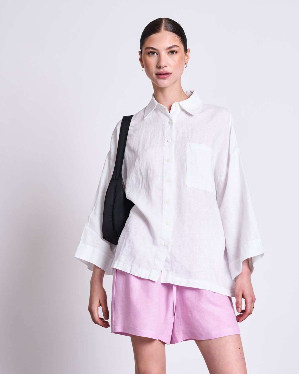 A woman stands against a plain background wearing the jan-n-june LINEN SHIRT MONS WHITE. She pairs the loose, classic-collar shirt with light pink shorts and a black shoulder bag, her hair tied back and her expression relaxed and confident.
