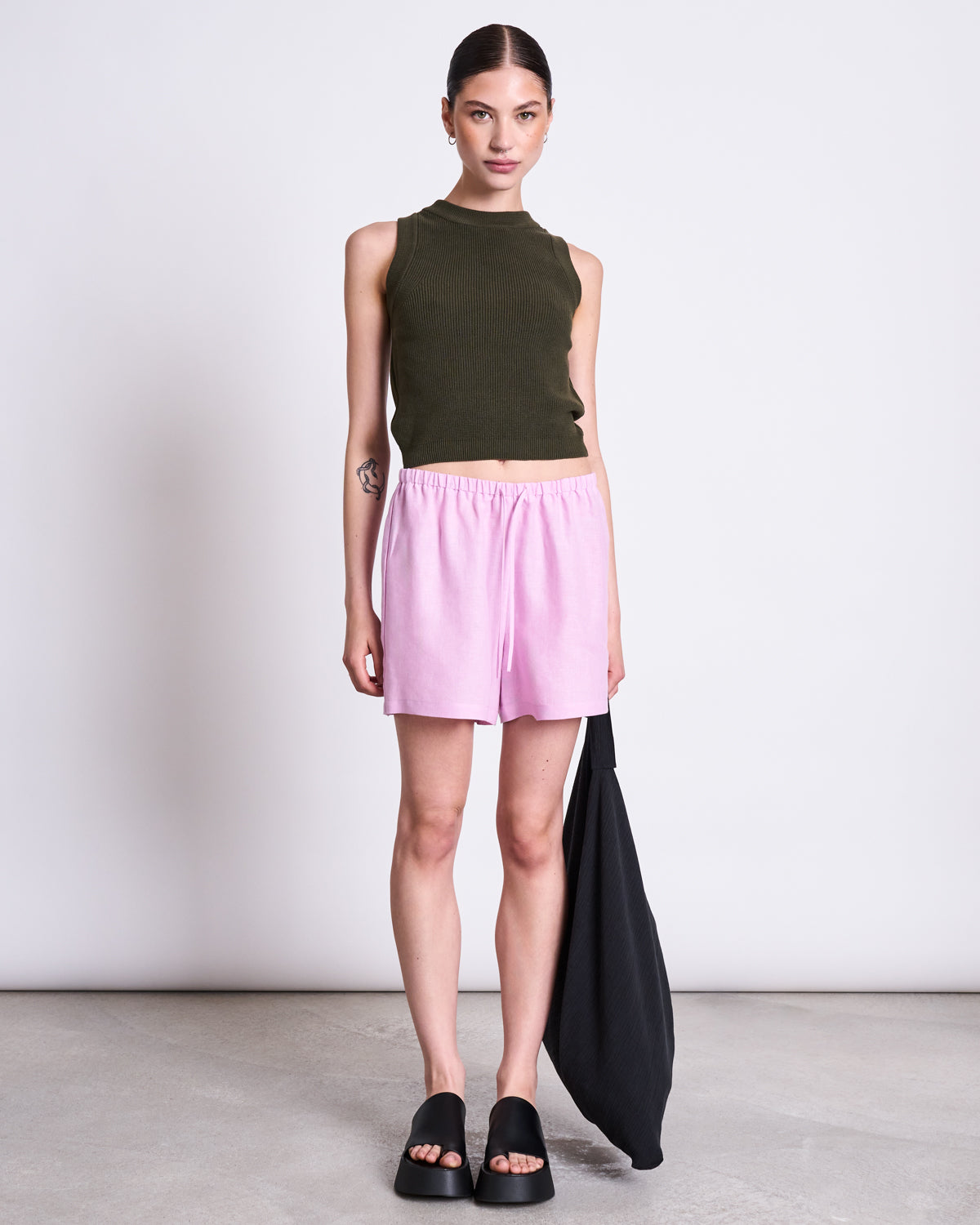 A woman stands against a white wall wearing the jan-n-june KNIT TOP GARDA DARK OLIVE GOTS, paired with light pink shorts and black slide sandals. She holds a black bag at her side and has her hair pulled back.