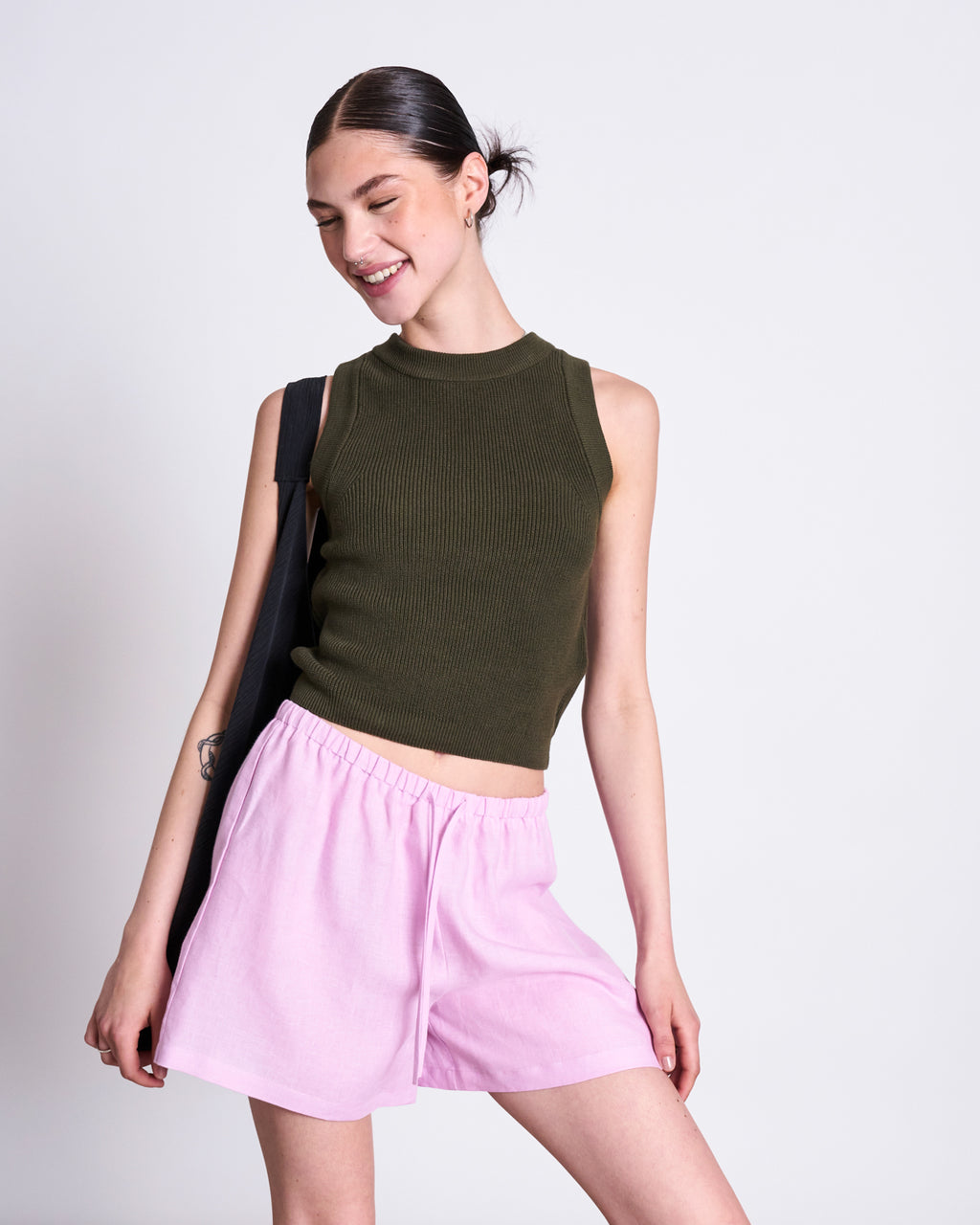 A smiling woman with dark hair in a bun wears the jan-n-june KNIT TOP GARDA DARK OLIVE GOTS, paired with light pink shorts and a black shoulder bag, posing against a plain white background.