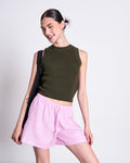 A smiling woman with dark hair in a bun wears the jan-n-june KNIT TOP GARDA DARK OLIVE GOTS, paired with light pink shorts and a black shoulder bag, posing against a plain white background.