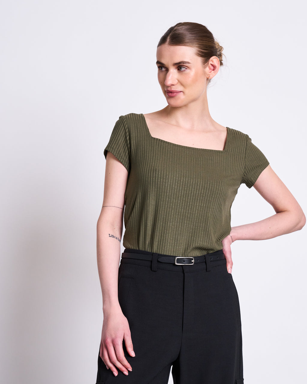 A woman stands against a white background wearing the jan-n-june T-SHIRT ARIMA DARK OLIVE, a short-sleeved, square neck top in soft Tencel™ rib fabric, paired with black high-waisted pants. Her hair is tied back and one hand rests on her hip.