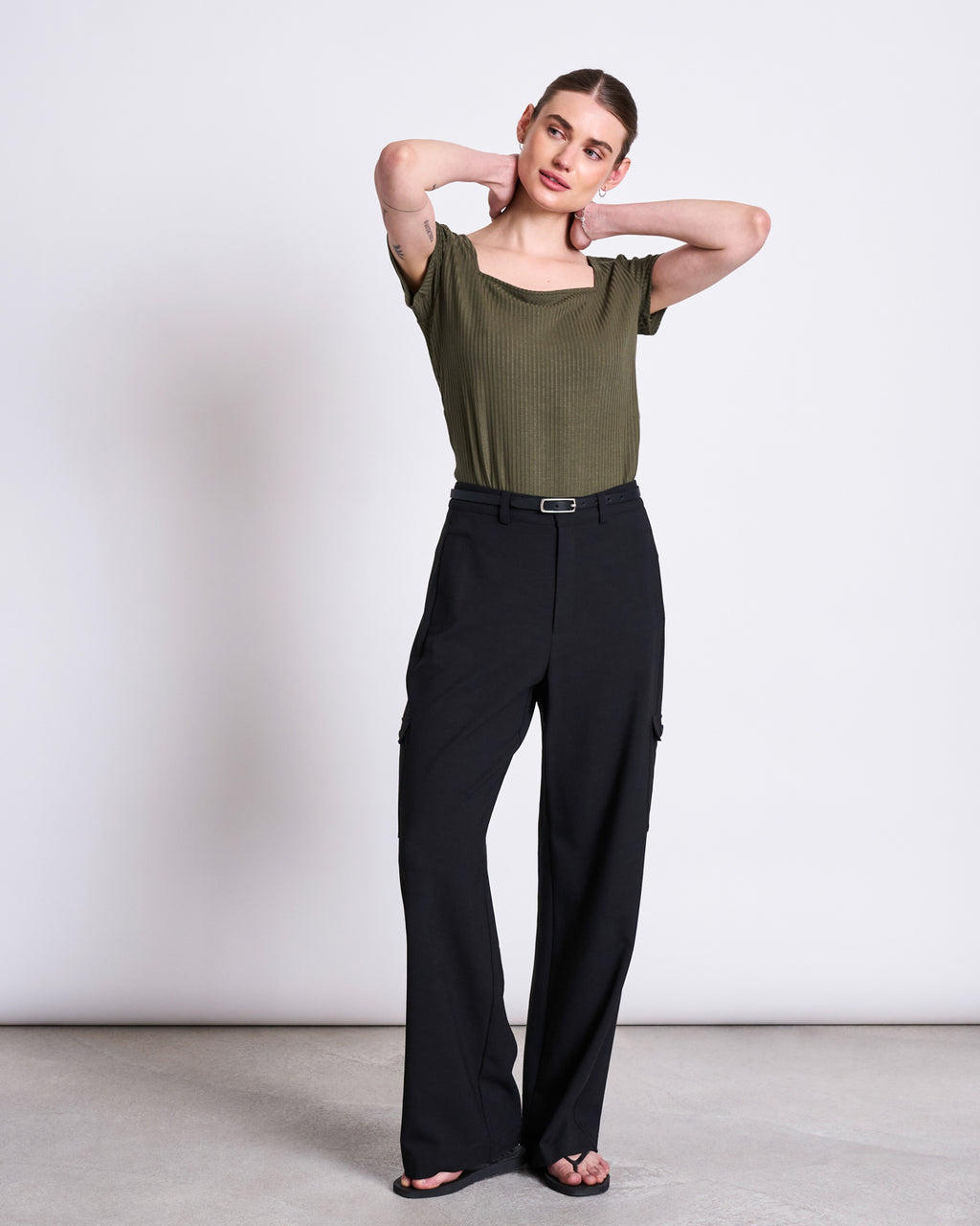 A woman poses against a plain background in the jan-n-june T-SHIRT ARIMA DARK OLIVE, a square neck short-sleeve top, paired with high-waisted black wide-leg belted pants. She stands with her hands behind her neck, gazing slightly to the side.