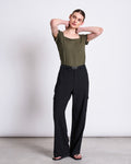 A woman poses against a plain background in the jan-n-june T-SHIRT ARIMA DARK OLIVE, a square neck short-sleeve top, paired with high-waisted black wide-leg belted pants. She stands with her hands behind her neck, gazing slightly to the side.