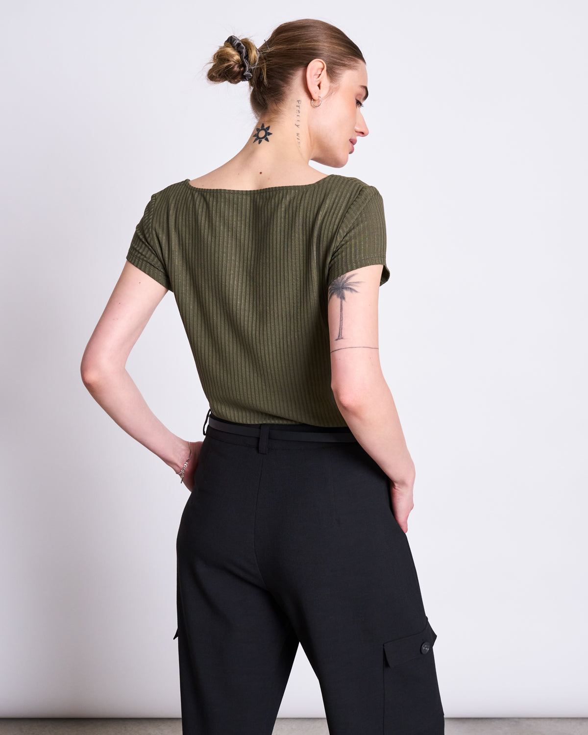 A person with their hair tied up and tattoos on their neck, arm, and behind the ear wears the jan-n-june T-SHIRT ARIMA DARK OLIVE, a soft square neck top in Tencel™ rib fabric, paired with black pants against a plain light gray background.