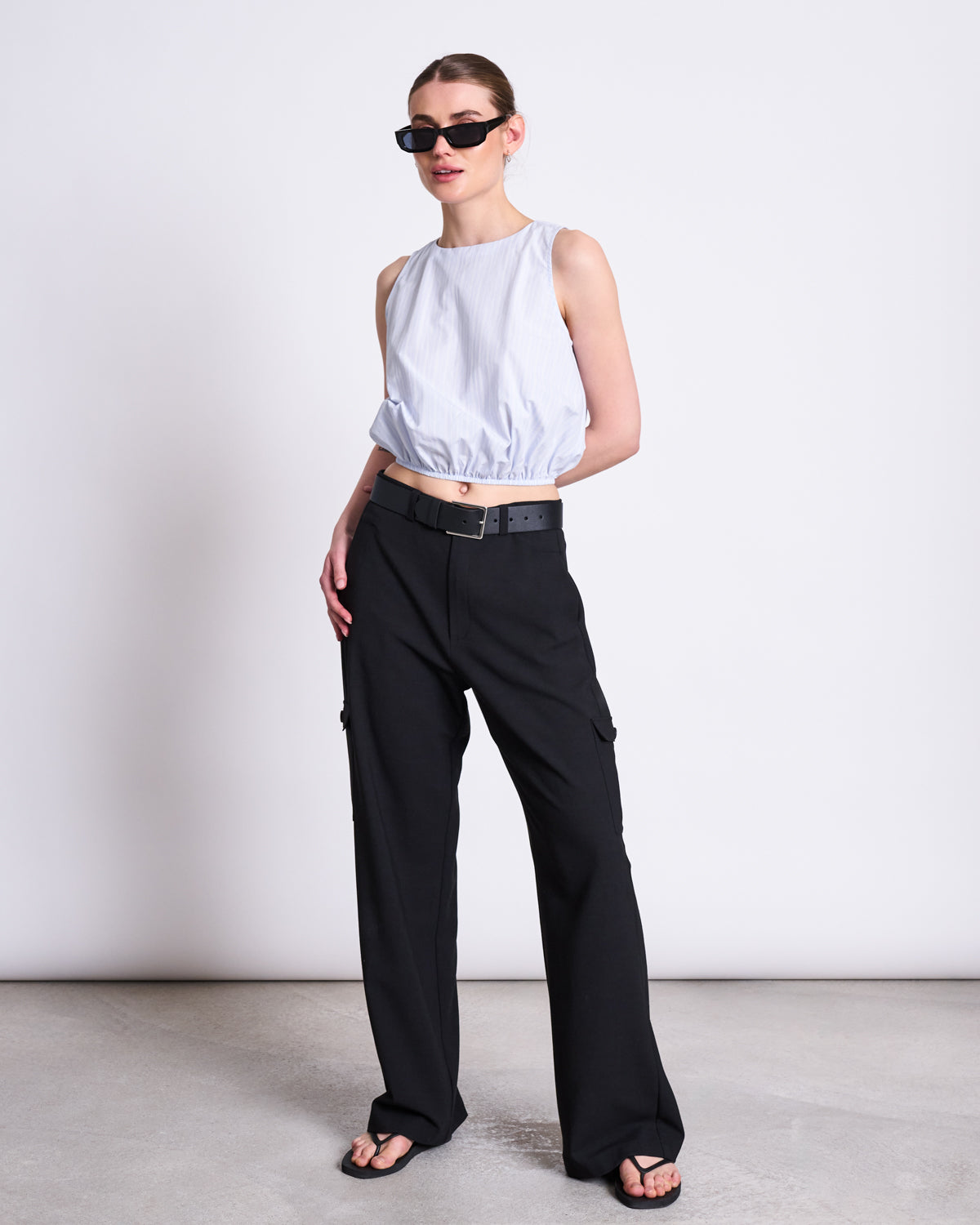 A woman in black sunglasses, a sleeveless light blue top, black wide-leg pants with the jan-n-june BELT FILSO BLACK SILVER, and black sandals stands confidently against a plain white background.
