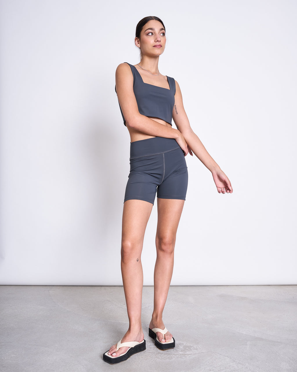 A woman wears jan-n-june ACTIVE BIKER SHORTS CRAIL SOLID GREY and a matching recycled polyamide crop top in a minimalist room, paired with white flip-flops. Her hair is tied back as she looks to the side with a neutral expression.