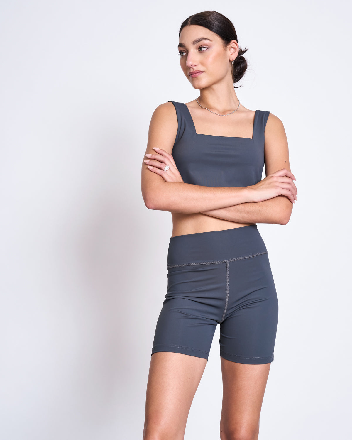 A woman stands against a plain background, wearing jan-n-junes ACTIVE BIKER SHORTS CRAIL SOLID GREY with a matching crop top in neoprene-like fabric. Her arms are crossed and she looks to the side with a neutral expression.