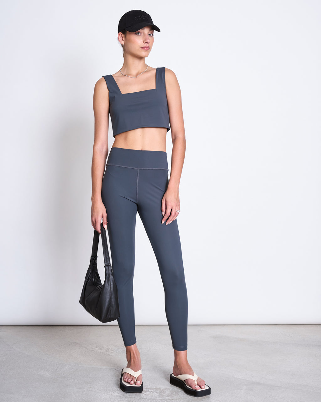 A woman in jan-n-june ACTIVE LEGGINGS HALLIN SOLID GREY made from recycled polyamide stands by a plain wall, paired with a dark blue crop top, black cap, thong sandals, and holding a black handbag.