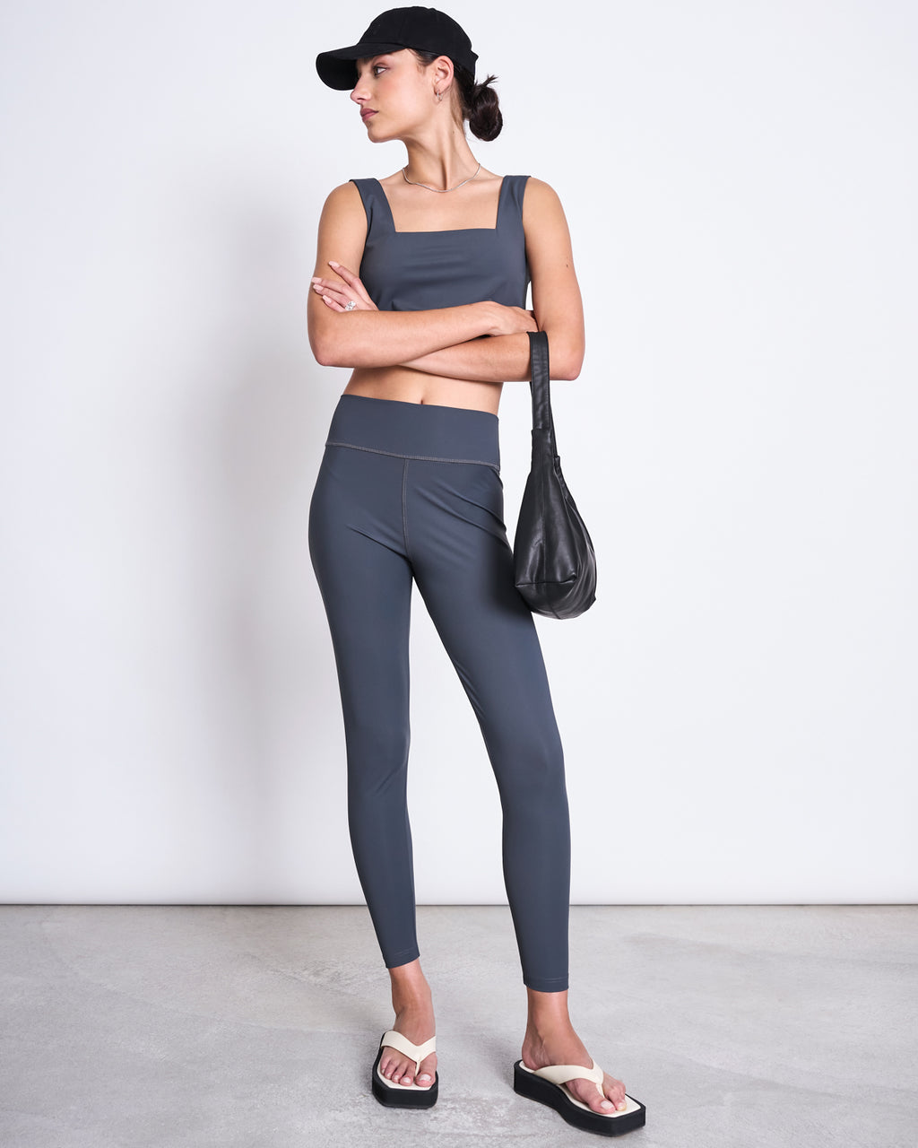 A woman stands against a white wall, wearing jan-n-junes ACTIVE LEGGINGS HALLIN SOLID GREY made from recycled polyamide with a neoprene-like feel, styled with a grey crop top, black cap, white flip-flops, and holding a black handbag.