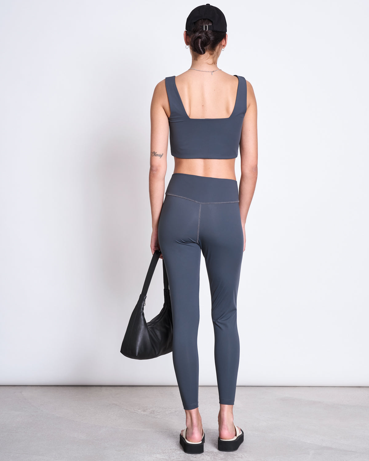 A person stands facing away, wearing jan-n-june ACTIVE LEGGINGS HALLIN SOLID GREY and a matching sports bra made from recycled polyamide resembling neoprene, styled with black sandals, a black cap, and a black bag against a plain white background.