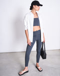 A woman in jan-n-june ACTIVE LEGGINGS HALLIN SOLID GREY, a black cap, white button-down shirt, gray crop top, and black flip-flops stands in a minimal studio with a black bag, looking to the side.