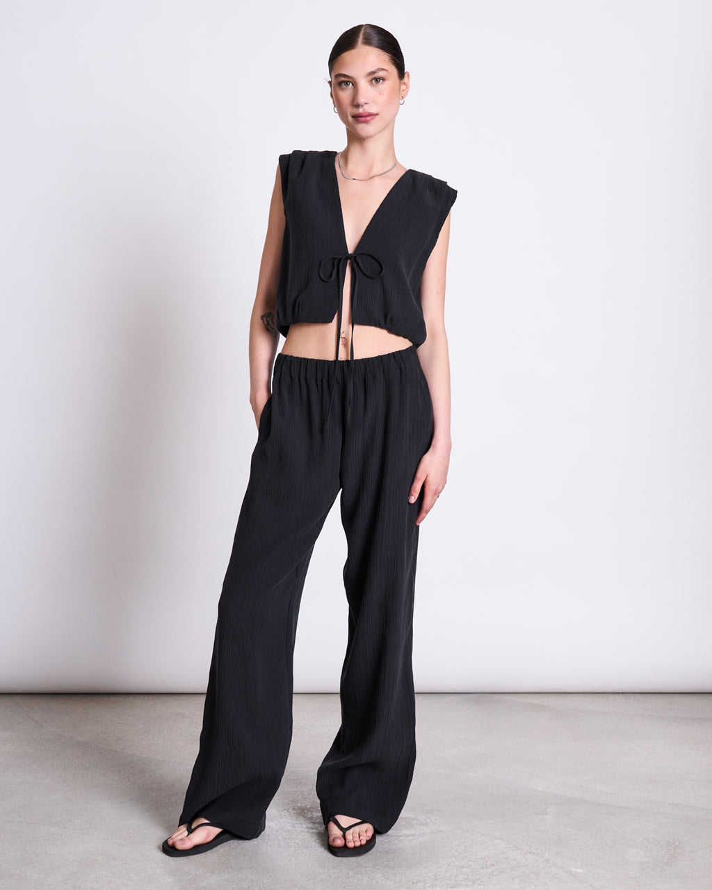 A woman stands against a plain background wearing the jan-n-june CRINKLE TIE TOP STROGET BLACK and loose-fitting black Tencel™ Lyocell pants, with her hands in her pockets and black sandals.