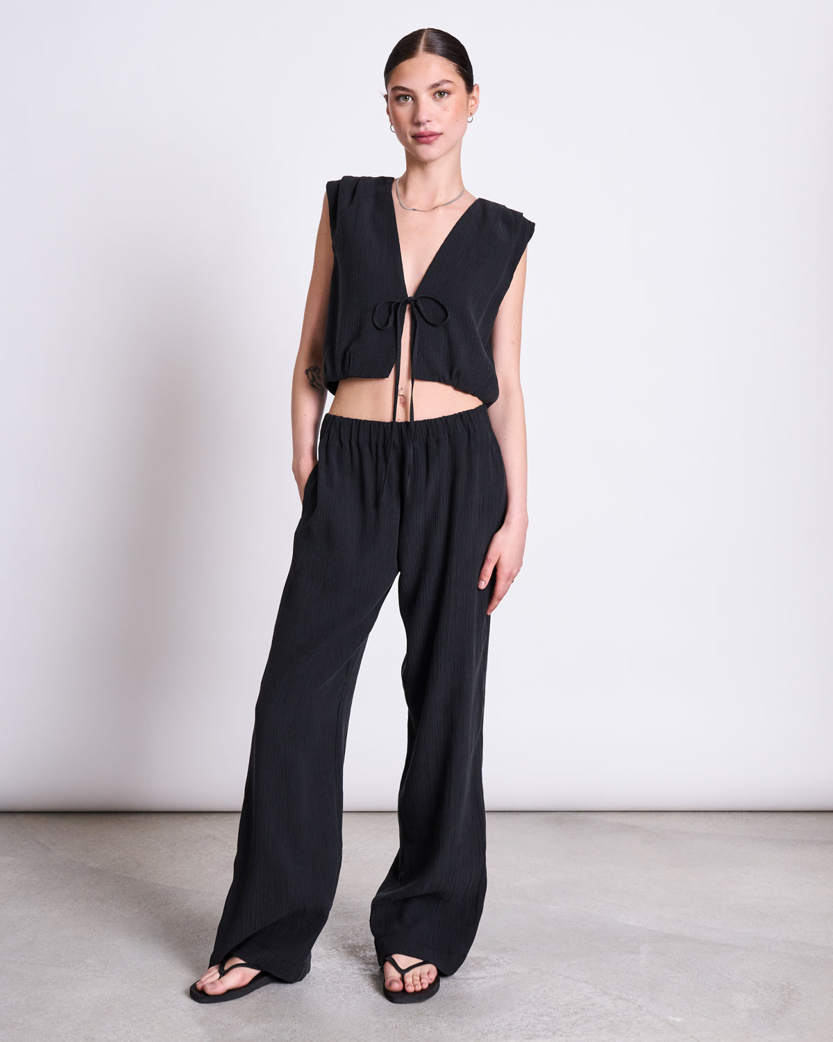 A woman stands against a plain background wearing a sleeveless, deep V-neck black crop top with front tie detail and jan-n-june CRINKLE PANTS HAVA BLACK made from soft Tencel™ Lyocell, her hands in her pockets and black sandals on her feet.