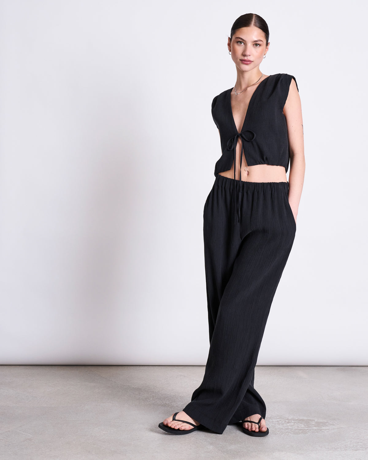 A woman poses against a plain background wearing jan-n-junes CRINKLE PANTS HAVA BLACK with a sleeveless black front-tie crop top, hair pulled back, black sandals, and one hand in her pocket.