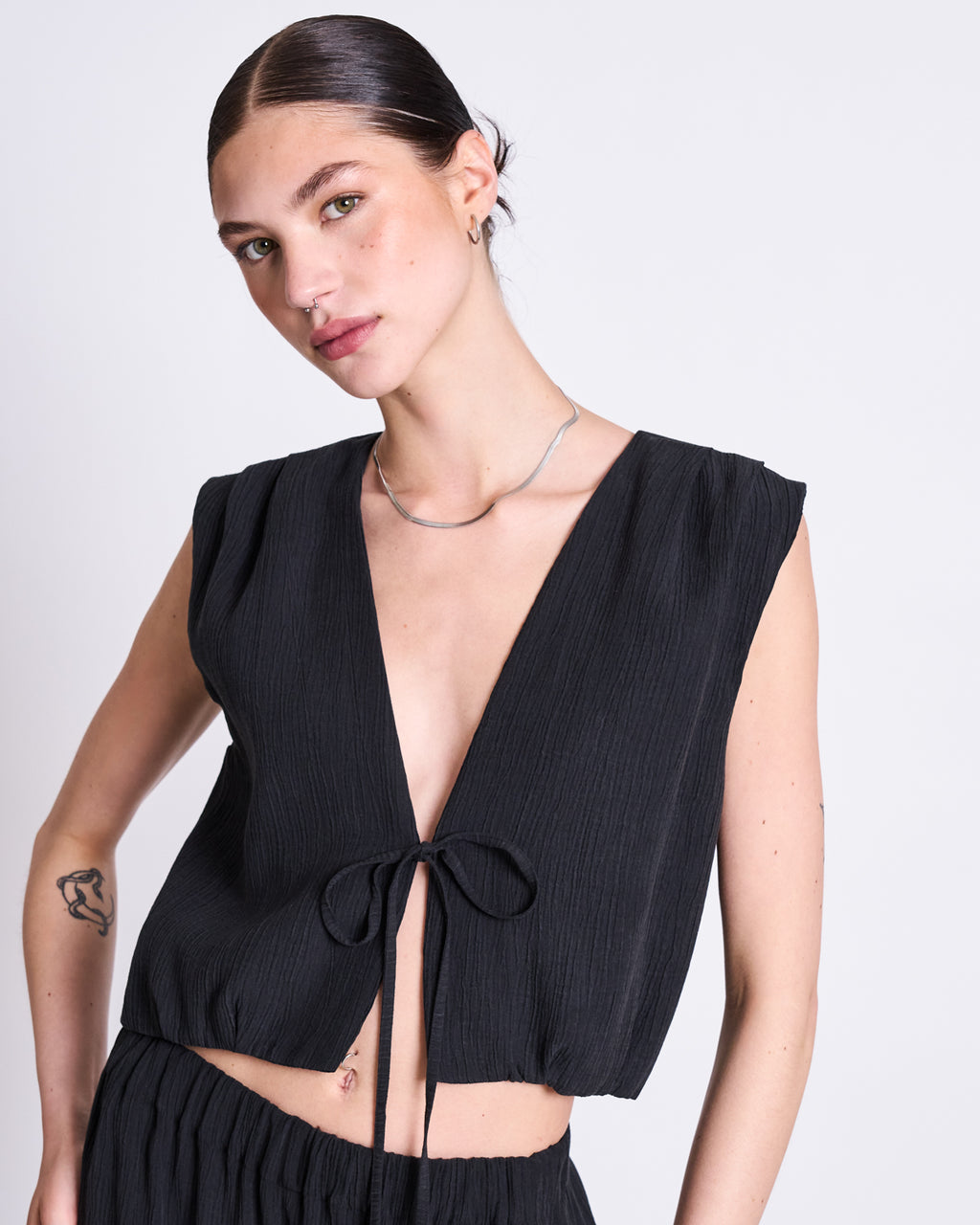 A woman with dark hair slicked back, wearing the jan-n-june CRINKLE TIE TOP STROGET BLACK—a sleeveless, deep V-neck top—paired with a silver necklace, poses against a plain light background. She has a tattoo on her right forearm.