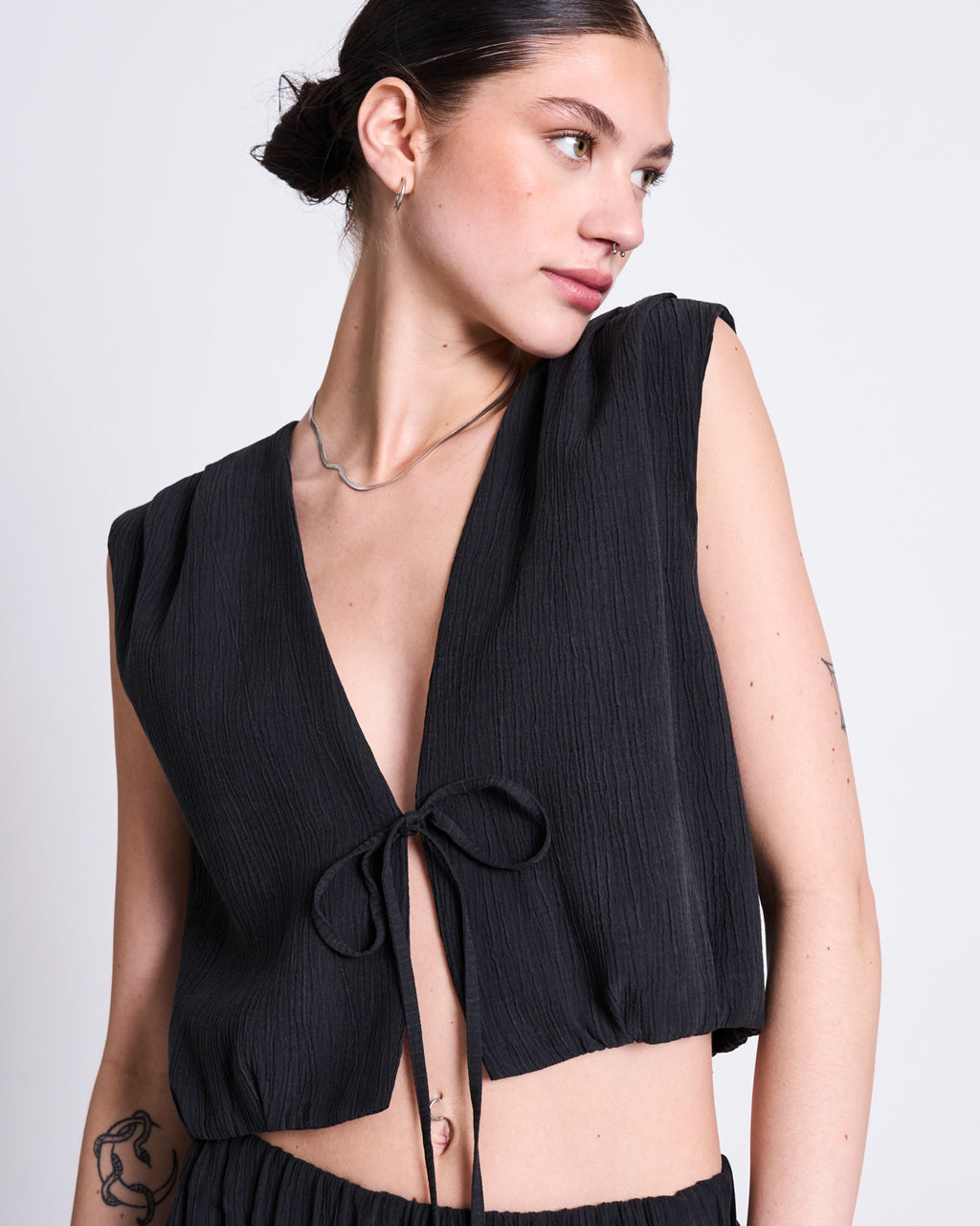 A person with dark hair tied back wears the jan-n-june CRINKLE TIE TOP STROGET BLACK, a sleeveless Tencel™ Lyocell top with a deep V-neck and front bow tie. A tattoo is visible on their arm. The background is plain white.