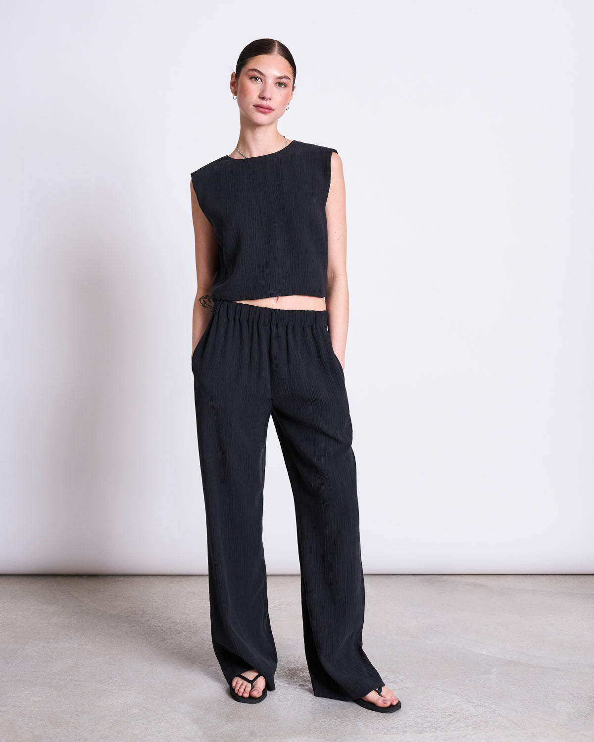 A woman stands against a white wall wearing the jan-n-june CRINKLE TIE TOP STROGET BLACK and matching loose Tencel™ Lyocell pants, with her left hand in her pocket and black sandals on her feet.