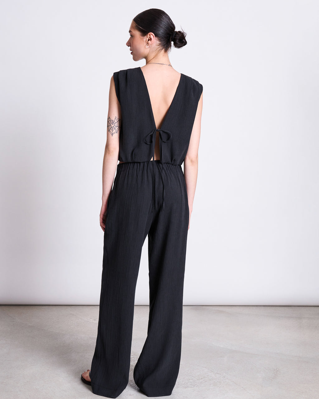A woman stands on STROGET wearing the jan-n-june CRINKLE TIE TOP in black, featuring Tencel™ Lyocell fabric, a deep V-back, and a lower back tie. Her dark hair is in a bun, revealing a geometric tattoo on her upper left arm.