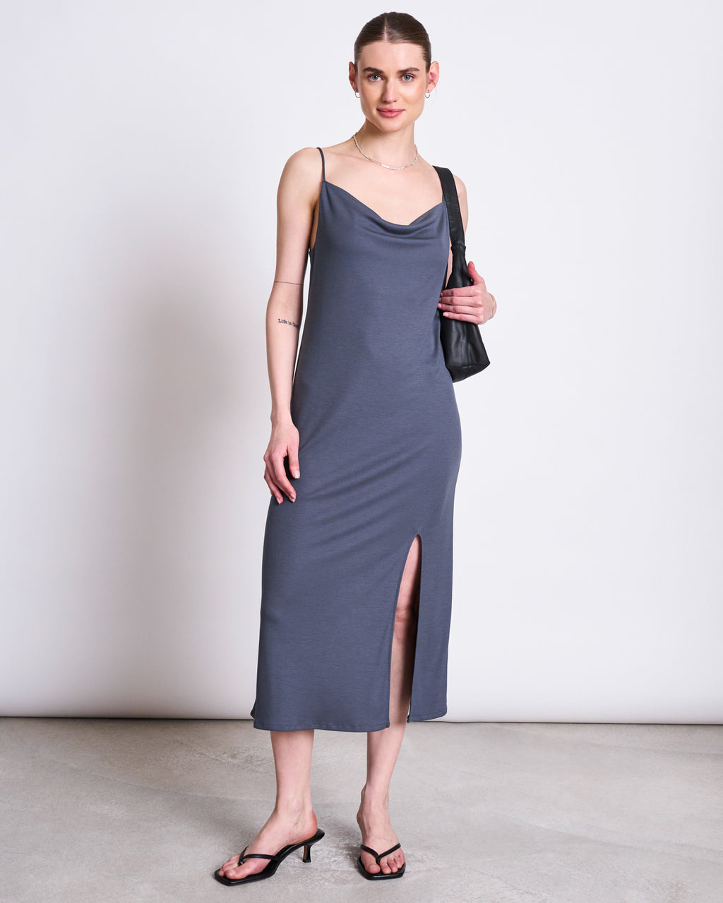 A woman smiles at the camera, wearing the jan-n-june MIDI DRESS HELEN TURBULENCE in gray with a cowl neckline, sleeveless design, front slit, plus black heeled sandals, a black shoulder bag, and hoop earrings against a plain background.