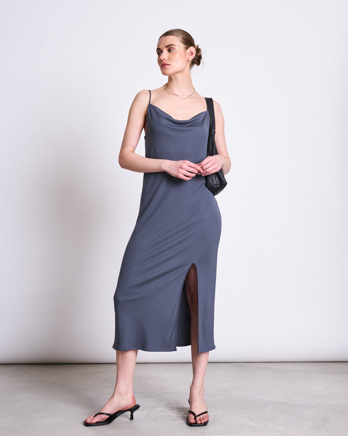 A woman stands against a plain background wearing the jan-n-june MIDI DRESS HELEN TURBULENCE, styled with black heeled sandals, a necklace, and a black shoulder bag. She looks to the side with her hands clasped.