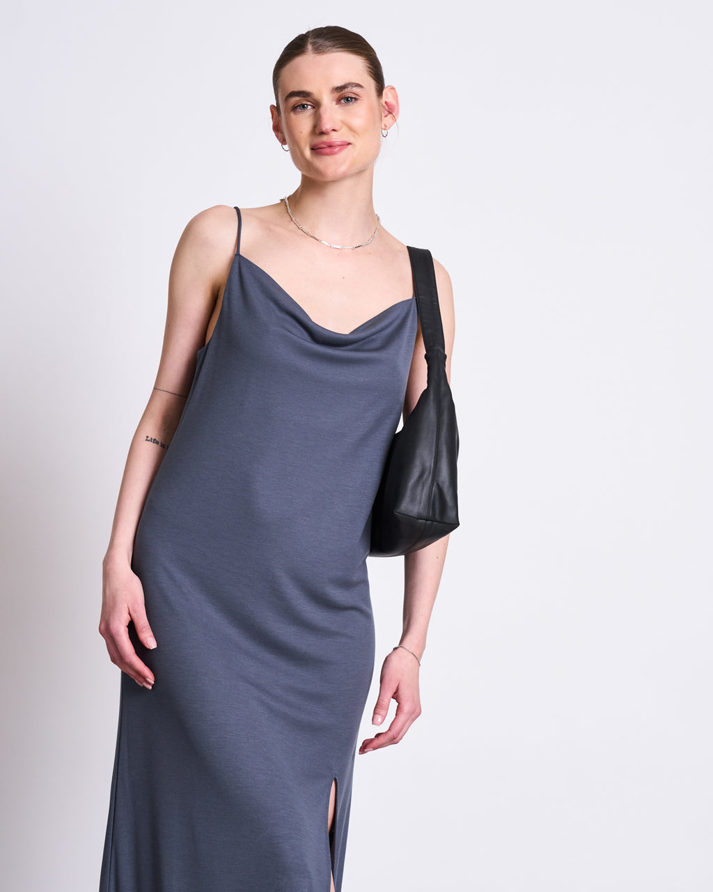 A woman smiles while wearing the jan-n-june MIDI DRESS HELEN TURBULENCE—a sleeveless gray midi dress with a side slit—and carries a black shoulder bag against a plain background.