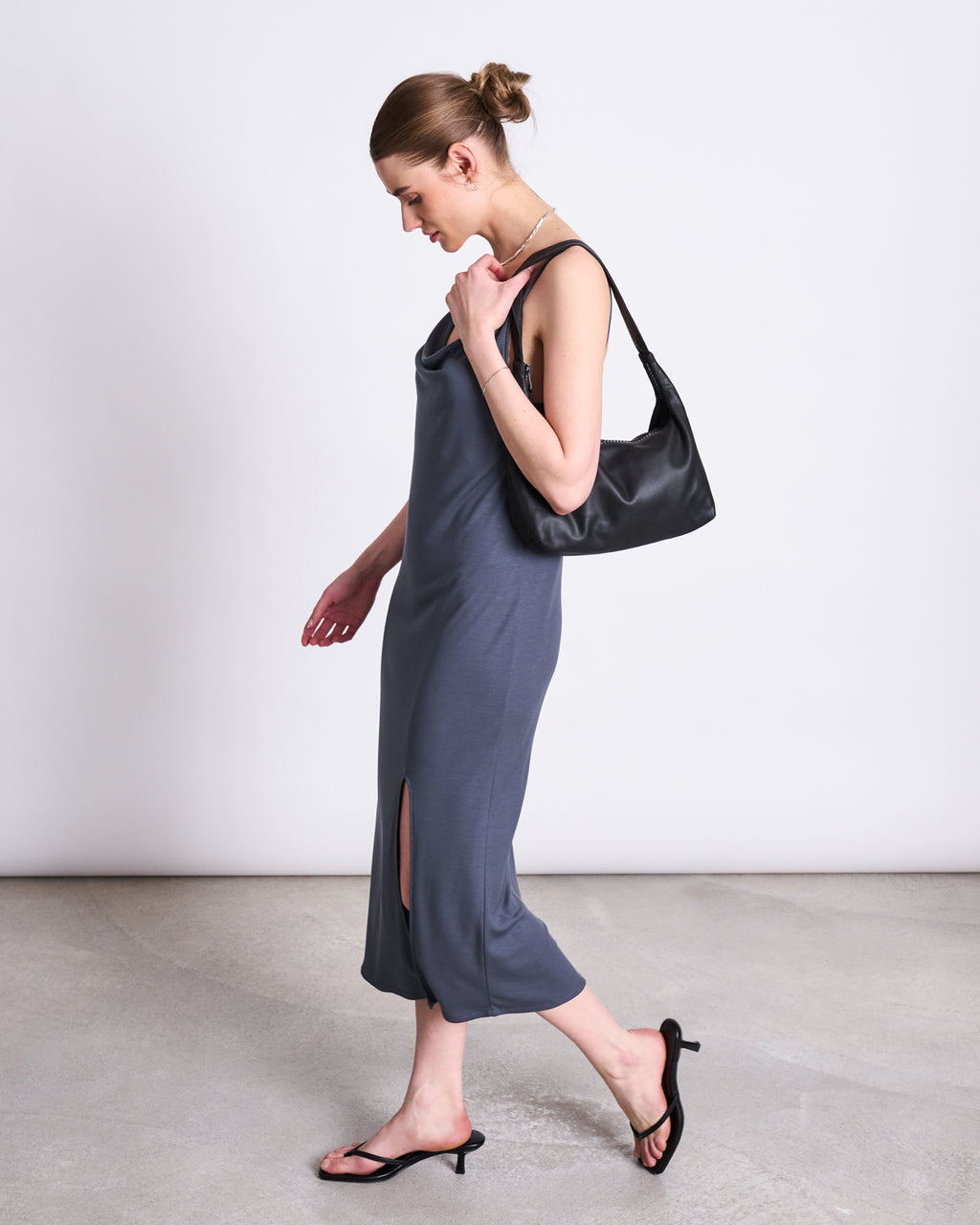 A woman wears the jan-n-june MIDI DRESS HELEN TURBULENCE in gray, styled with a black shoulder bag and black heeled sandals. Her hair is in a bun as she walks in profile, smiling slightly against a plain background.