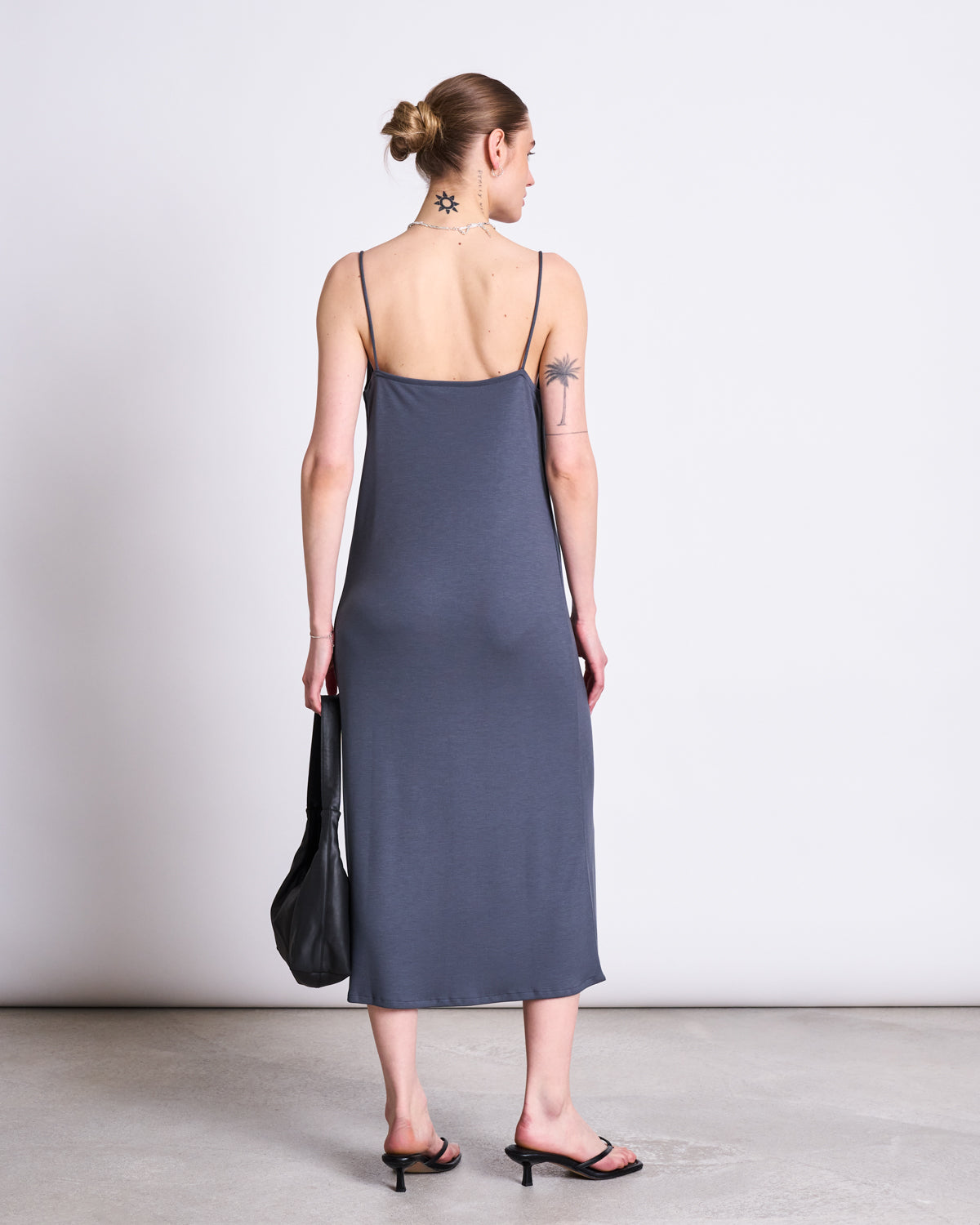 A person with a low bun and tattoos faces away, wearing the jan-n-june MIDI DRESS HELEN TURBULENCE— a sleeveless gray sustainable slip dress—paired with black heeled sandals and a black bag, set against a plain background.