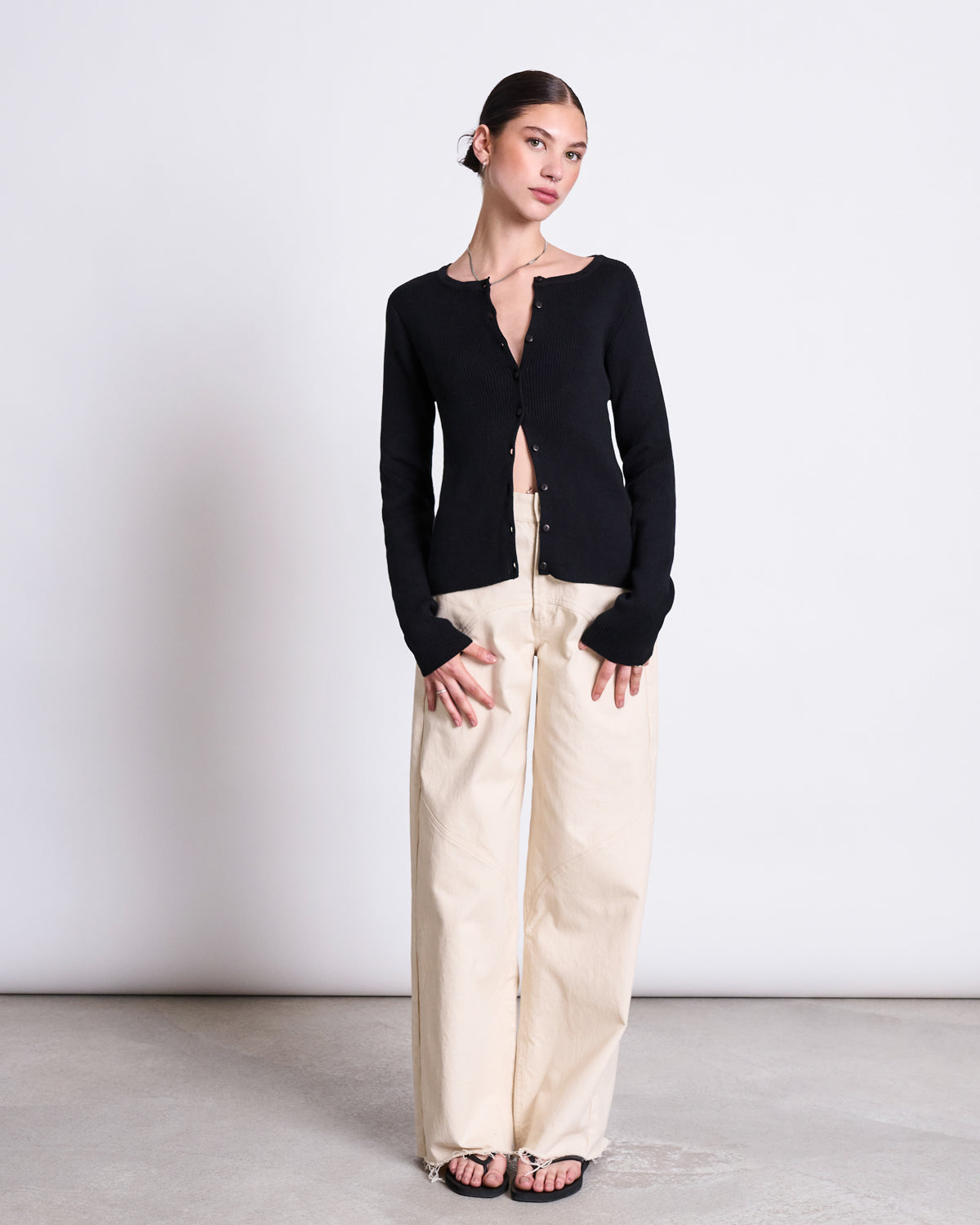 A woman stands against a plain background wearing the jan-n-june CREWNECK CARDIGAN GLIMT BLACK GOTS, paired with cream wide-leg pants and black sandals. Her hair is tied back and her hands rest by her sides.