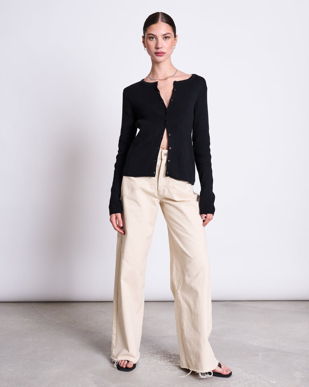 A woman stands against a plain backdrop wearing the jan-n-june CREWNECK CARDIGAN GLIMT BLACK GOTS, cream wide-leg pants, and black sandals. Her hair is pulled back and she gazes at the camera with a neutral expression.
